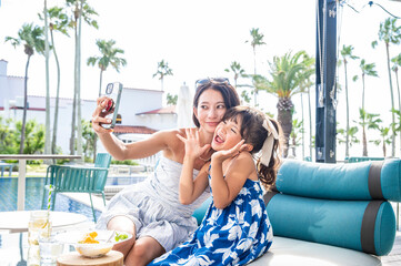 Japanese mother and daughter taking a fun selfie together with a smartphone at a tropical resort poolside café during summer vacation with palm trees sunshine and playful family travel atmosphere