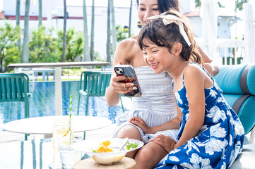 Japanese mother and daughter sitting together at a resort poolside café looking at a smartphone and laughing during summer vacation with palm trees sunshine and tropical travel atmosphere