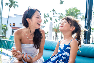 Japanese mother and daughter laughing together at a resort poolside café during summer vacation with palm trees bright sunlight and joyful tropical family travel atmosphere