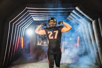 American football player wearing uniform and helmet emerging from a stadium tunnel, flexing biceps with smoke and light effects
