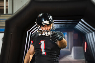 American football player in uniform and helmet emerging from a stadium tunnel, pointing at viewer with confidence and focus