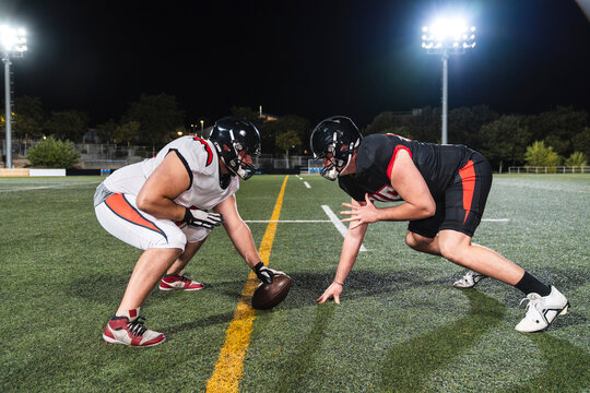 Two american football players wearing helmets and uniforms, crouching on a green field under stadium lights, ready for action - Powered by Adobe
