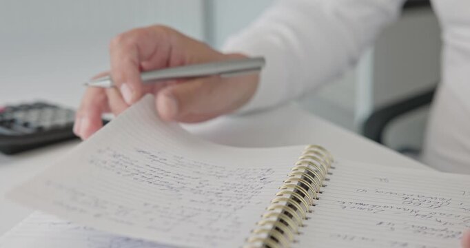 A close-up of a man in a shirt checking notes and making corrections with a pen in a notepad on a metal spring on an office desk