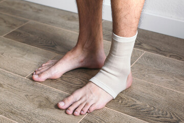 Close-up of male feet on wooden floor, one ankle wrapped in elastic bandage support. Concept of...