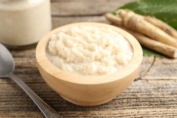 Tasty horseradish sauce, spoon and roots on wooden table, closeup