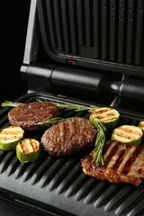 Electric grill with beef steaks, zucchini and rosemary on black table, closeup