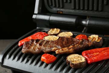 Electric grill with beef steaks and vegetables on grey table, closeup
