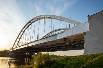Sunset view over a river with a modern steel arch bridge and city skyline in the background. Astana, Kazakhstan, Argar Bridge over the Ishim River