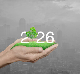 2026 white text with growing tree on green grass field in hands over pollution of city tower and skyscraper, Happy new year 2026 ecological cover concept