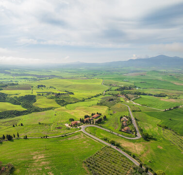 Aerial view of rolling green hills and winding roads converge near rustic buildings, the landscape stretching towards distant mountains, Pienza, Tuscany, Italy.