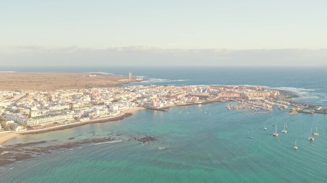 Drone sunrise shot over Playa del Medio Beach Area in Fuerteventura, Canary Islands, Spain, revealing a coastal town, marina, and sunlit Atlantic shoreline.