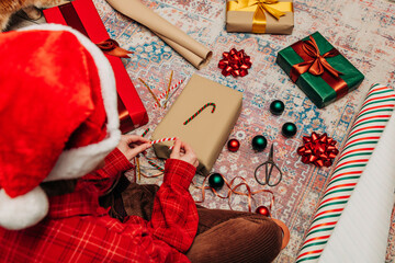 woman in hat sits on cozy rug surrounded gift boxes, wrapping Christmas presents