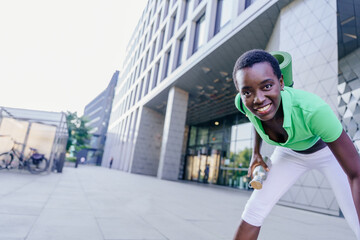 Young African woman with yoga mat and bottle smiling in front of modern glass building