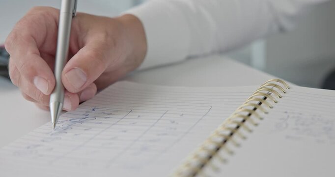 A close-up of a man in a shirt checking notes and making corrections with a pen in a notepad on a metal spring on an office desk