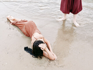 Female figure lies partially submerged on wet beach sand, head resting on arm, waves washing around her body while a second figure stands barefoot nearby in shallow water.