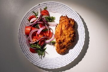 A golden-brown fried chicken breast is served on a decorative plate with a tomato salad consisting of tomatoes, red onions, and greens.