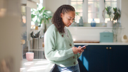 Teenage Girl At Home In Kitchen Connecting With Friends On Social Media Using Mobile Phone
