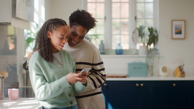 Mature Mother With Teenage Daughter At Home In Kitchen Looking At Mobile Phone