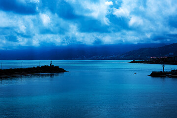 A dramatic coastal scene with dark blue waters under heavy clouds. The silhouettes of mountains and city buildings line the horizon. While a lighthouse and a bird add detail to the moody seascape.