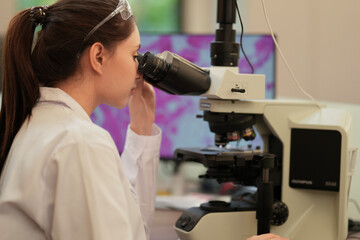 A female scientist in a white lab coat and safety goggles meticulously analyzes data on a computer screen, pointing at a key finding during her research in a modern laboratory.