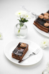 Rich chocolate cake slice on plate with fork and white flowers clean bright background.