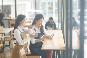 Two Asian female waitress wearing apron using digital tablet taking orders from customer in cafe, SME entrepreneur seller business concept