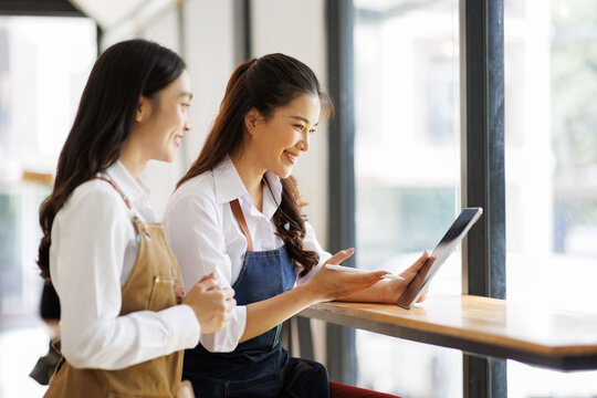 Two Asian female waitress wearing apron using digital tablet taking orders from customer in cafe, SME entrepreneur seller business concept
