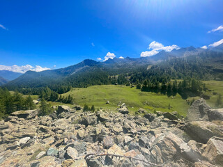 Rugged alpine terrain in the Mont Avic Natural Park, located in the Aosta Valley, Italy. The foreground features jagged rocks and sparse vegetation, including coniferous trees and small bushes.