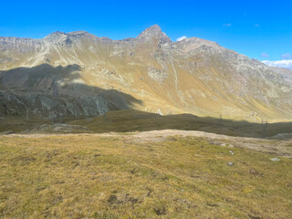 Expansive mountain landscape in the Mont Avic Natural Park, located in the Aosta Valley, Italy. The foreground features a grassy plateau with patches of bare earth, while dramatic rocky peaks rise