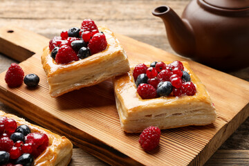 Tasty puff pastries with berries and teapot on wooden table, closeup
