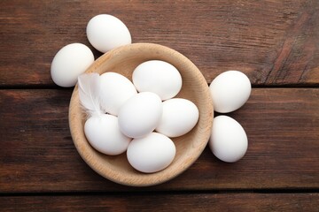 Raw chicken eggs in bowl and feather on wooden table, flat lay