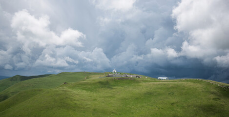 Remote observatory buildings atop a green hill under a dramatic, cloud-filled sky, casting shadows and light across the rolling landscape, evoking a sense of isolation and the scale of nature