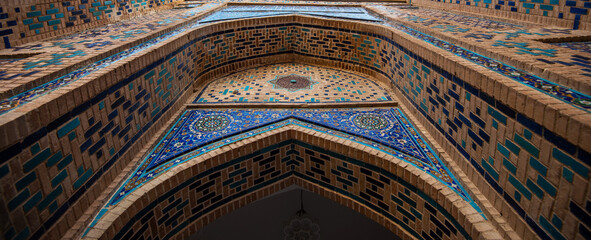 Ornate Islamic architecture with blue mosaic tile work and detailed geometric patterns on a brick facade, photographed from below within the historical necropolis complex in Samarkand, Uzbekistan.
