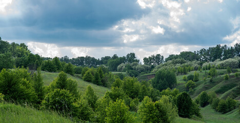 Rolling green hills speckled with trees under a dramatic sky, where shafts of sunlight pierce through clouds, presenting a dynamic and serene rural landscape.