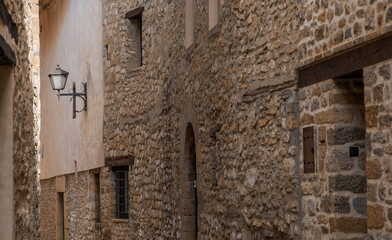 A narrow alleyway in a historic Spanish village, flanked by rustic stone walls and old buildings, with a charming wrought-iron lamp and small barred windows.
