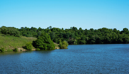 serene landscape features a lush, green lakeside surrounded by rolling hills and a variety of trees under a clear blue sky. the tranquility and natural beauty of a pristine rural area