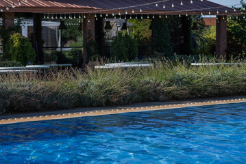 poolside relaxation area with a covered outdoor seating space, lush greenery, and string lights hanging above, featuring a blue water pool, wooden benches, and potted plants in a peaceful garden