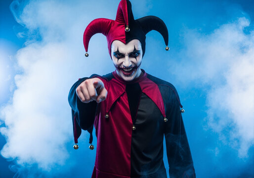 Scary evil jester in a Halloween costume pointing at the camera. Sinister man with horror makeup and a menacing grin on a smoky blue background.