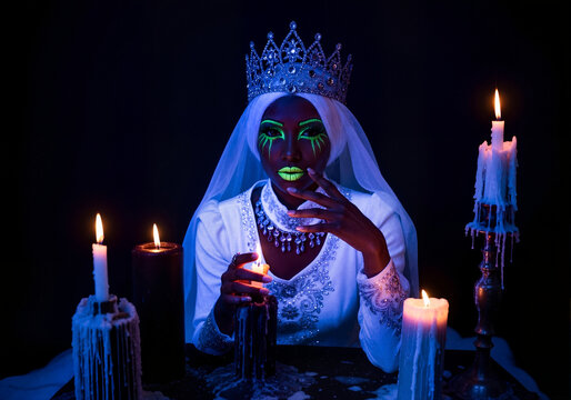A mystical queen with glowing neon green UV makeup for Halloween. Fantasy portrait of a woman with a crown surrounded by candles in the dark. Supernatural magic and ritual concept