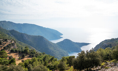 Breathtaking panoramic view of a serene Mediterranean coastline with lush greenery, overlooking a calm bay under the soft light of a summer afternoon. The rugged beauty of a mountainous region leading