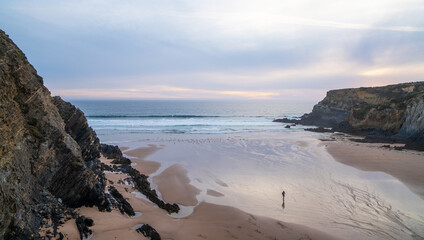 A peaceful coastal scene at sunset with a solitary figure walking on the sandy beach. The waves...