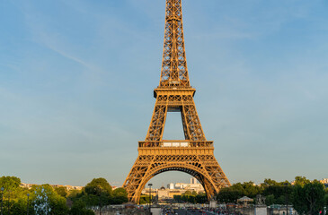 Eiffel Tower at sunset with clear skies, surrounded by green trees and city architecture, Paris, France. Iconic landmark and symbol of Paris, captured in warm evening light.