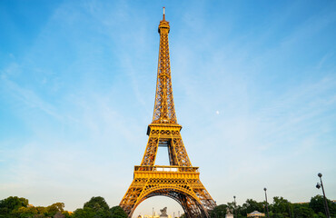 Eiffel Tower in Paris illuminated by soft evening light, framed by water fountains on either side, with the moon visible in the clear blue sky, creating a classic Parisian scene.