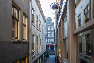 Narrow pedestrian alleyway with traditional tall buildings and vintage lanterns in central Amsterdam, Netherlands. The passage leads to a wider street with bicycles and a canal in the distance.