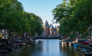The Waag, a medieval city gate turned museum and restaurant, stands at the end of a canal in Amsterdam, Netherlands, framed by green trees, moored boats, and parked cars on a summer day.