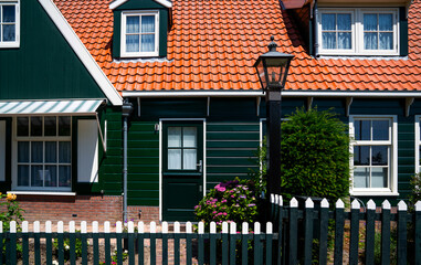 green wooden house with a red tiled roof and white-framed windows in a traditional european village, featuring a black lamppost, a white picket fence, and blooming hydrangea flowers near the entrance