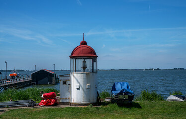 Small vintage lighthouse with a red dome and white base stands on a grassy shore by the Markermeer lake in Marken, North Holland. Sailboats, a pier, and boat storage structures are visible nearby.