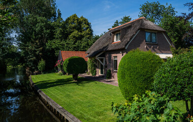 Traditional brick house with thatched roof in Giethoorn, Netherlands, surrounded by neatly trimmed topiary, red-tiled shed, flowering plants, and green lawn under a clear blue summer sky.