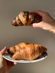 A fresh golden croissant served on a white plate held in human hands against a neutral background.