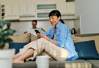 Portrait of an asian woman and her husband, a couple, chatting on phone at home, using modern mobile phone and working on laptop, checking social media
